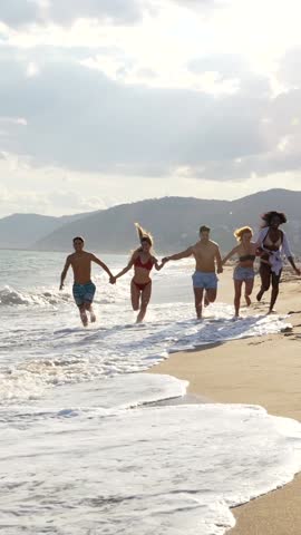 Multicultural friends running and holding hands together along sunny shoreline, embodying joy and freedom of summer beach vacation