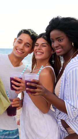Young friends capturing vibrant moments while enjoying tropical beach party, drinking cocktails and taking spontaneous group selfies with diverse companions