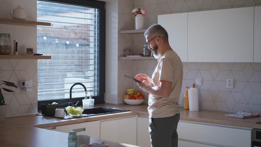 Mature man adjusting windows blind in the kitchen by tablet.