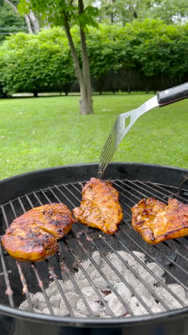 Grilling Meat on a Charcoal Grill. Hot charcoal grill with three chicken breasts cooking on top. A man uses a spatula to adjust the placement of the chicken. Backyard setting.
