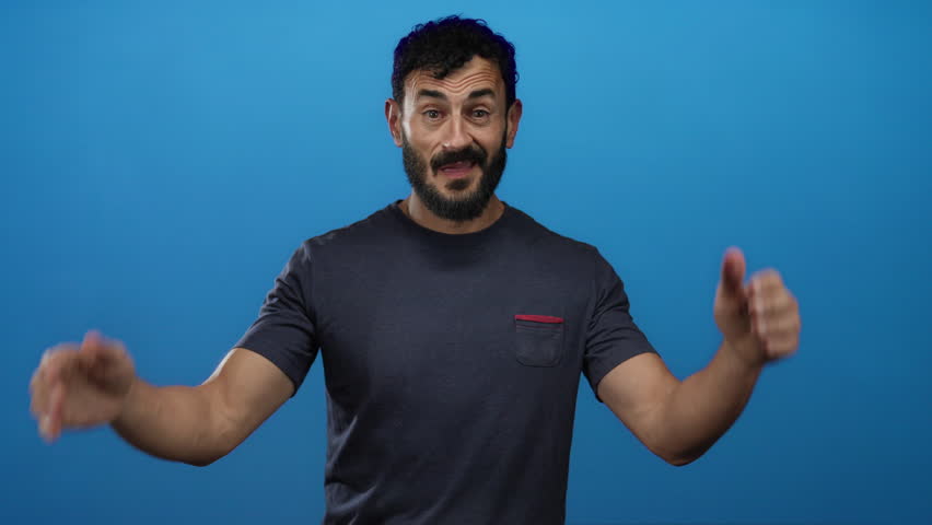 Hispanic man with beard gesturing happily in front of a vibrant blue background, wearing a casual dark t-shirt, expressing joy and openness with wide hand movements.