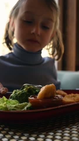 Little girl enjoys golden nugget with sauce, broccoli and lettuce on plate, cheerful healthy eating scene in bright family cafe.