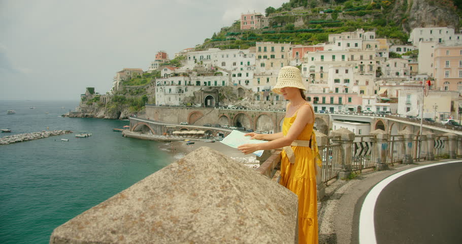 Woman studies a map on a coastal road overlooking an Italian town Atrani. Tourist admires architecture from scenic viewpoint on Amalfi Coast, Italy, sunny day