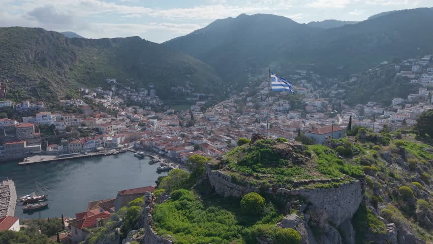 A calm morning view of Hydra’s traditional port, seen from the hilltop with the Greek flag waving in the foreground. Peaceful light and island beauty in one frame