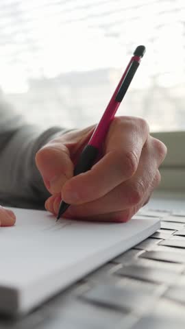 Left-handed man writing on paper next to a window