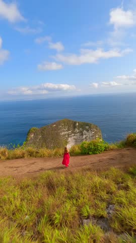 Cinematic FPV drone shot of woman in red dress walking toward dramatic cliffs and turquoise waters at famous Kelingking Beach on Nusa Penida island, Bali, Indonesia