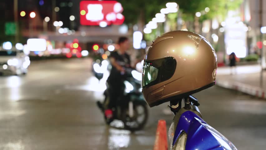 Close-up of a motorcycle helmet mounted on a rearview mirror of parked taxi motorbike beside a brightly lit city road at night. Copy space safe motorcycle riding rules and tips concept