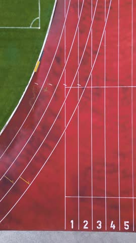 Aerial view of a red running track with marked lanes and curved lines. Athletic stadium section with part of a green football field. Sports and fitness concept.