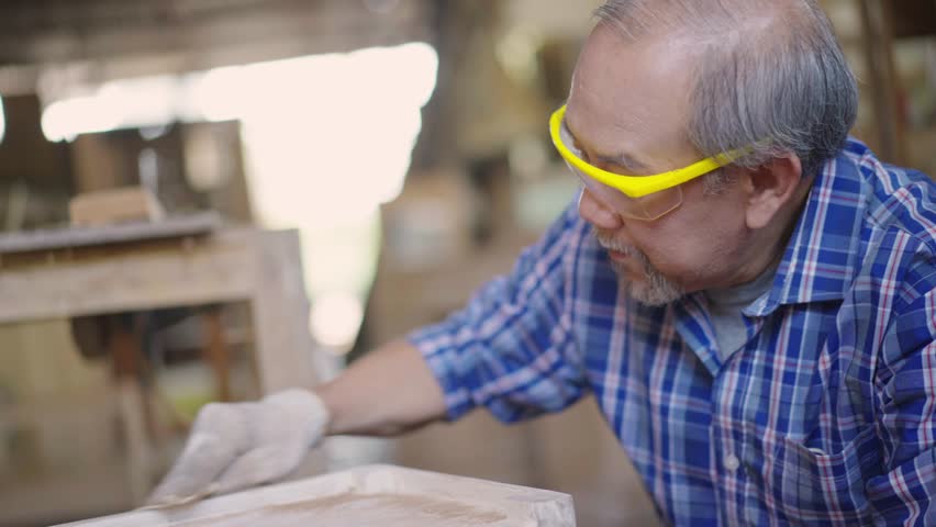 An elderly carpenter is using sandpaper to smooth the surface of wooden furniture in a wood workshop. The image showcases craftsmanship, attention to detail, and the traditional woodworking process
