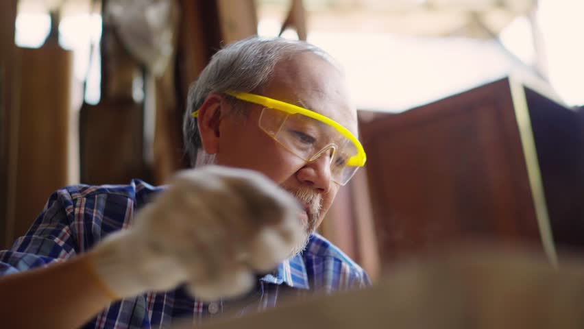 An elderly Asian male carpenter sanding a wooden piece of furniture in a carpentry workshop. He is focused on his craftsmanship, working carefully with sandpaper to smooth the wood surface