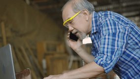 An elderly carpenter is multitasking in his wood workshop, checking customer orders on a laptop while speaking on the phone, reflecting small business operations and customer communication. - Powered by Shutterstock - Get 15% off with code: PIKWIZARD15