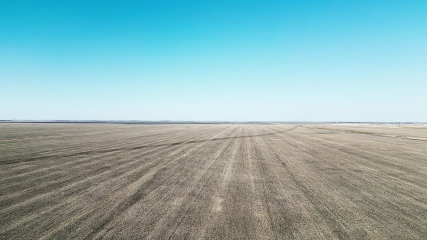 Drone floats over barren dirt field; horizon meets cloudless blue sky
