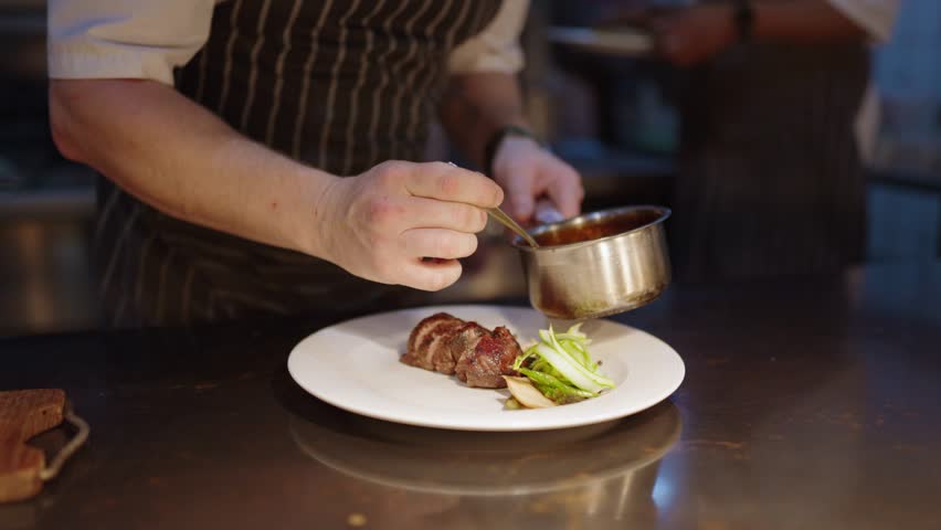 View of a chefs hand pouring sauce over a perfectly cooked piece of meat as he plates up a meal in a fine dining restaurant