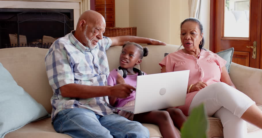 Diverse grandparents and granddaughter using laptop together on couch, enjoying family time, at home. bonding, technology, intergenerational, connection, computer, quality time