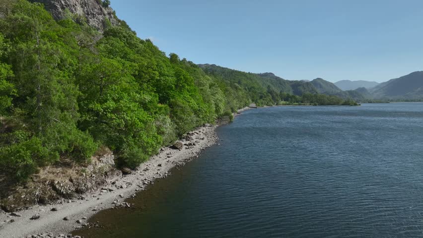 Wooded and craggy lake shoreline with mountains in the distance on sunny spring day. Derwentwater, Lake District, Cumbria, UK.