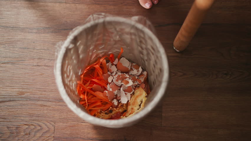 waste bin containing bright carrot peels broken egg shells and leftover snack on wooden floor with partial view of person disposing food waste into bin near chair