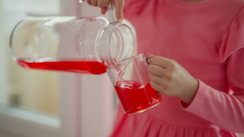 close up of little girl wearing pink dress pouring red juice from glass jar into clear mug spilling drink on cloth indoors with soft blur background playful cheerful