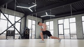 Young man stretching feet and legs with forward lunge in bright gym room, wearing white tank top and black shorts, large windows and equipment racks in background, showcasing warm up, and strength - Powered by Shutterstock - Get 15% off with code: PIKWIZARD15