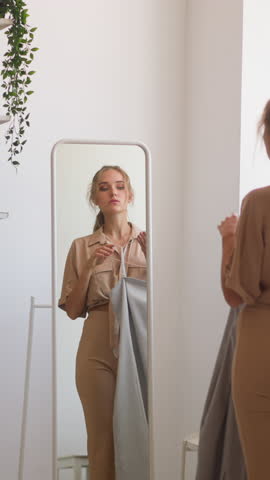 Blonde woman with ponytail holds elegant silver dress on hanger choosing feminine outfit for birthday celebration against mirror slow motion
