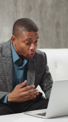 Positive African-American man tells funny story to colleagues at online meeting via modern laptop computer sitting at table in living room slow motion