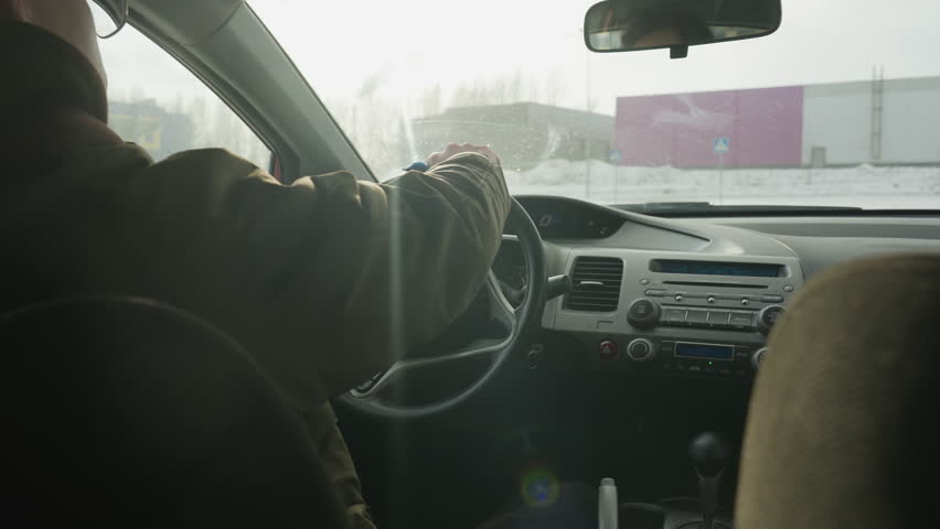 rear view of man cleaning steering wheel with blue cloth inside parked car during bright winter day showing dashboard, frosted window, and snowy outdoor environment through windshield
