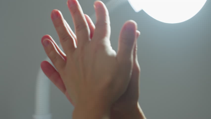 Wellness technician sprays hydrating mist onto hands then rubs palms together under bright clinical light, creating shimmering droplets for hygienic skin prep before facial treatment