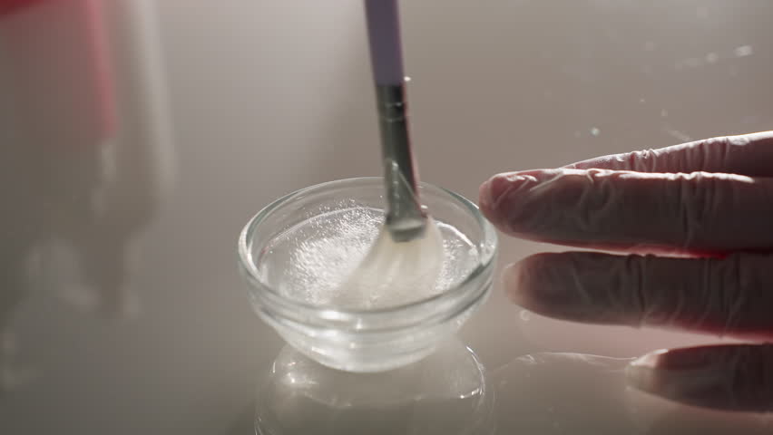 Hands in gloves mix creamy facial mask in small glass bowl with silicone brush on reflective spa trolley under soft light, conveying professional skincare ritual and hygienic precision care