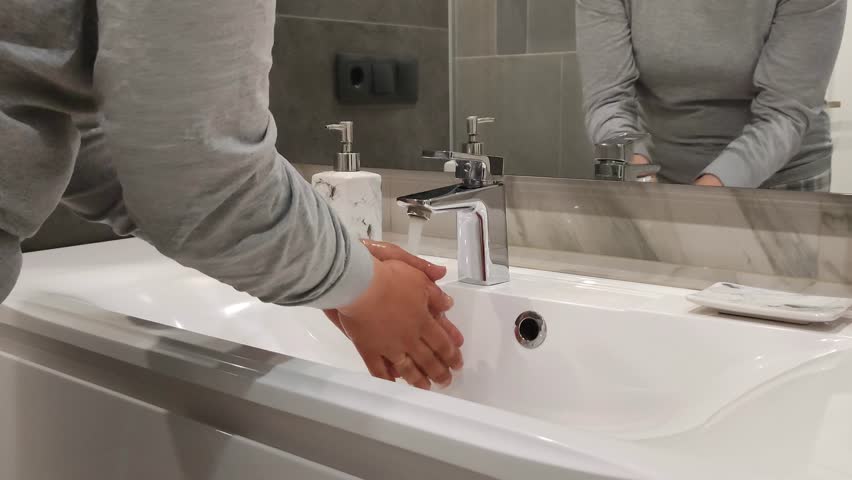Woman washes her hands with clean water under faucet and shakes water off her wet fingers. Hygiene and body care. Reflection of young woman in large toilet or bathroom mirror. High quality 4k footage