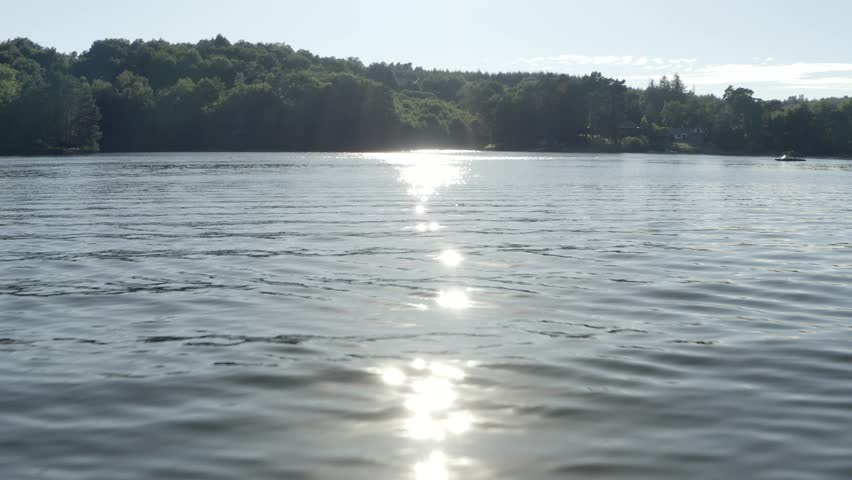 Aerial view flying low over rippling lake water surface towards sun glare and forested shore, Marcillac La Croisille, France