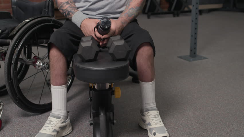 Sweaty bearded man with disability sitting on leather bench, drinking water and wiping sweat from forehead while taking rest after workout in gym - Powered by Shutterstock - Get 15% off with code: PIKWIZARD15