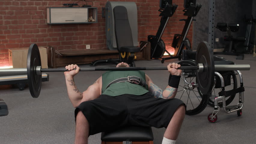 Unrecognizable man dressed in sportswear and weight lifting belt lying on bench while doing bench press with heavy barbell