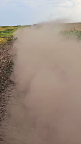 Car Kicking Up Dust on Dirt Road, White car speeds along a dirt road, creating a large cloud of dust surrounded by cornfields and sunlit countryside