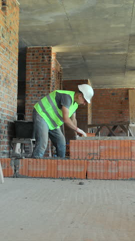 Bricklayer Working on Interior Wall Construction. A bricklayer, dressed in a high-visibility vest and hard hat, is seen working on an interior wall within a partially completed building.