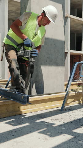 Construction Worker Drilling on Building Site. A construction worker is seen drilling into a concrete slab on a building site, wearing a high-visibility vest and hard hat.