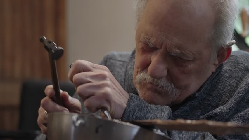 Elderly master craftsman on a wheelchair works in his workshop engraving horimono image on the blade of a Japanese sword with hammer and chisel