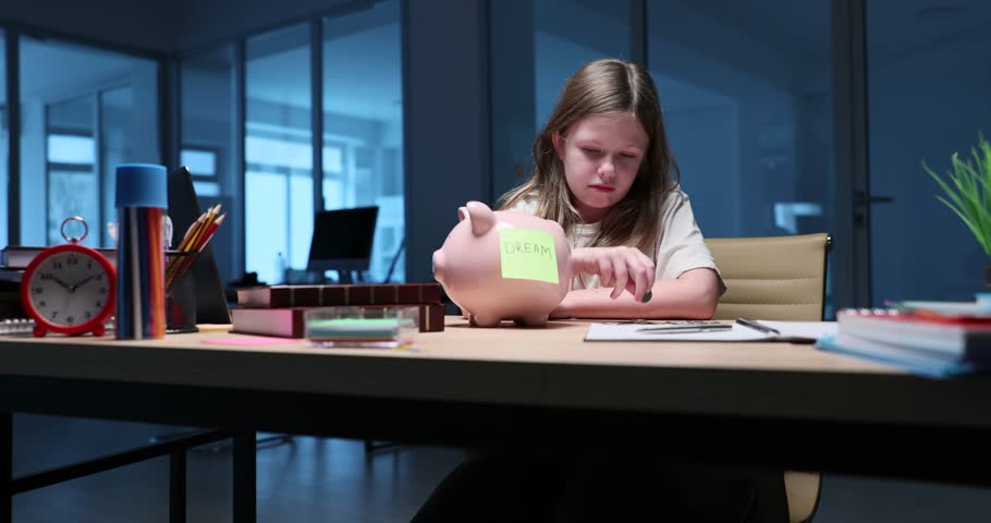 Concentrated young girl professional inserts coins into dream labeled piggy bank in minimalist workspace. Diligently saving for future aspirations