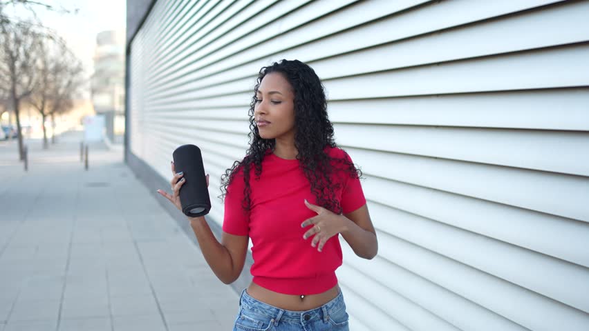 On a beautiful, sunny day, a fashionable young woman stands outdoors, confidently holding a portable speaker while joyfully enjoying her favorite music, radiating a sense of freedom and fun