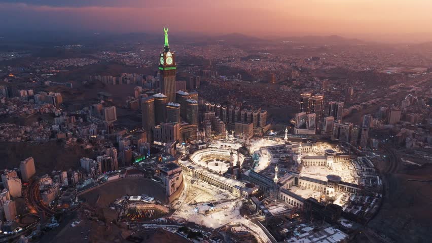 Aerial view of the Abraj Al-Bait Clock Tower and Grand Mosque in Mecca illuminated by the golden light of sunrise, surrounded by the sprawling holy city and mountains