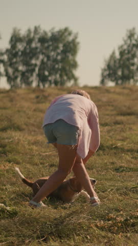 Animal instructor in pink shirt and shorts training dog to roll over on farmland, dog excitedly follows instruction with tongue out, bright sunny day, with background of distant trees