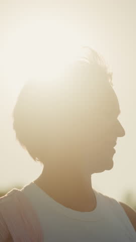 Woman standing outdoors with bright sunlight radiating behind her, smiling warmly while looking at something in distance, sunlight creates halo effect around her head