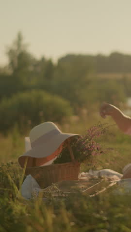 Woman in open grassy area under sunlight reaching for sun hat on woven basket filled with flowers, enjoying peaceful summer day outdoors on picnic mat, with blurred natural background