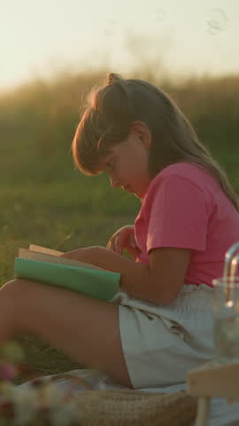 Little girl sitting outdoors during golden hour reading book and turning page, surrounded by picnic setup with fruits, croissant, and bubbles gently floating in air, sunlight illuminating background