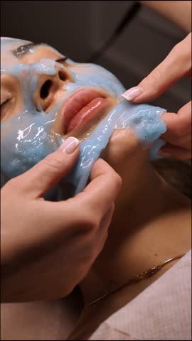Professional cosmetologist peeling off facial blue alginate mask from female face in beauty spa salon, healthcare and beauty. Beautician removes Hydrogel mask off woman's skin, anti-aging, vertical