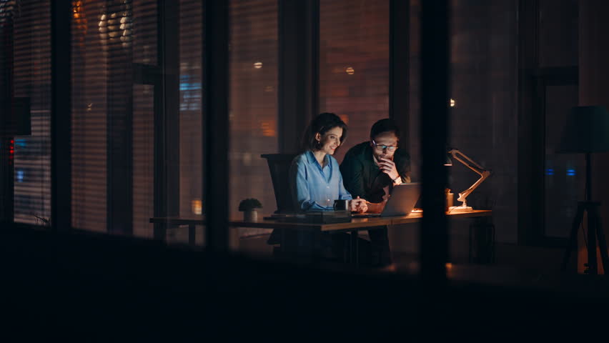 Hardworking businesspeople browsing laptop at dark office open space. Smiling businesswoman presenting online project to businessman at night. Happy colleagues working on company development overtime.