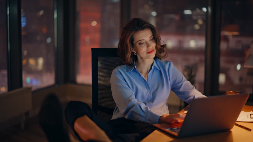 Elegant manager reading email at laptop sitting comfortably at office late evening closeup. Smiling businesswoman with legs on desk browsing computer at night. Successful startuper surfing internet.