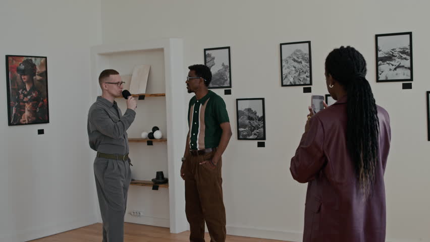 Interview in art gallery with young African American man speaking to microphone and woman recording video via smartphone