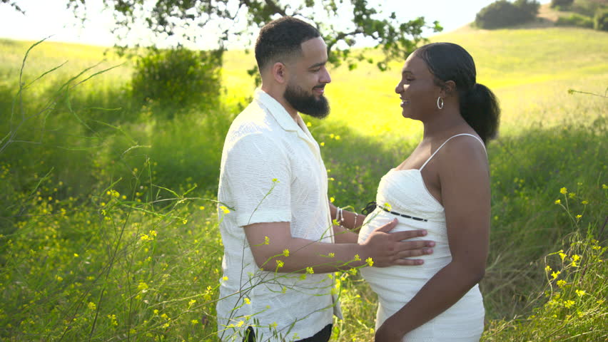 A joyful African American couple is celebrating their pregnancy outdoors amidst a beautiful lush summer landscape, fully embracing their love and the wonderful anticipation of welcoming new life soon