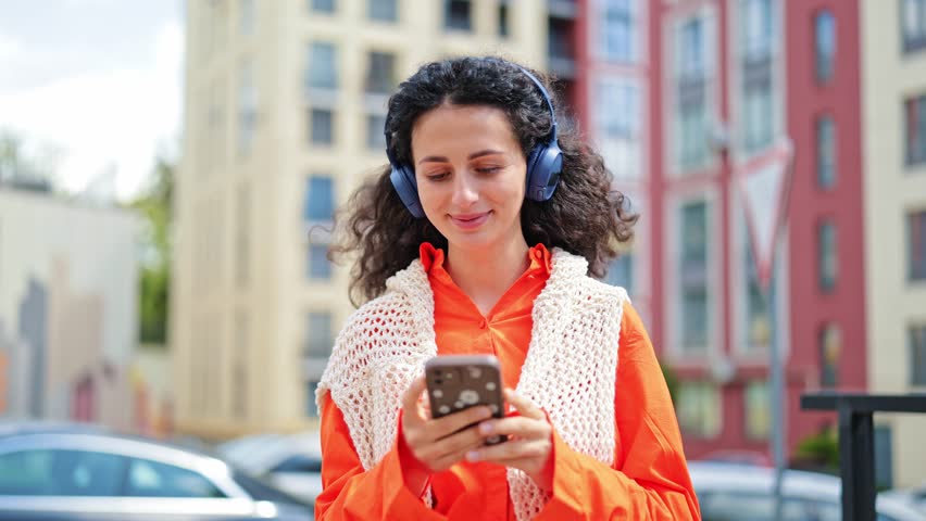 Beautiful cheerful woman in an orange shirt and white vest listens to music and using a phone on a city street.