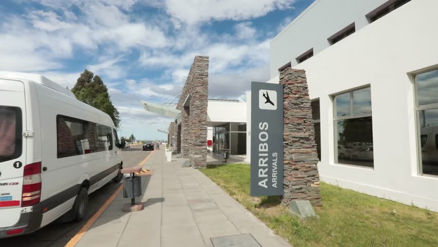 little airport entrance with arrivals, travelers wheeling luggage, welcoming signs, glass doors reflecting sunlight—evoking movement, anticipation, and the start of journeys.