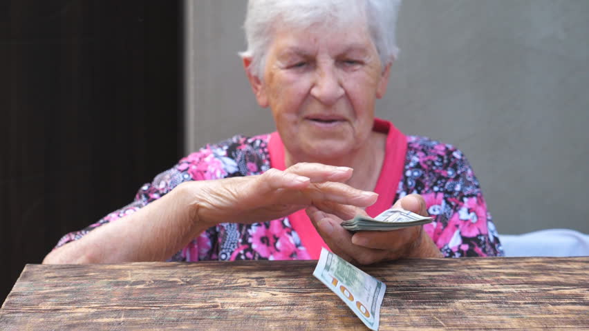 Portrait of happy grandmother holding bundle of money in hands and scattering foreign currency on the desk. Old smiling woman throwing one hundred dollar bills on the table. Close up Slow motion